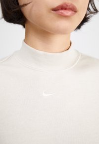 Šviesiai bežinis Nike mako kaklo džemperis su prigludusia forma, pagamintas iš minkštos, tekstūruotos medžiagos. Priekyje yra mažas baltas logotipas.