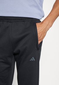 Svarta sportbyxor med elastisk midja, sidofickor och en grå Adidas-logotyp på nedre benet, med en slät, matt textur.