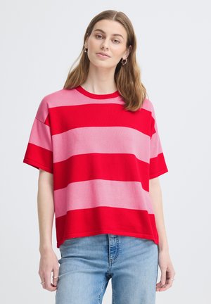 Jeune femme aux cheveux châtain clair portant des boucles d'oreilles argentées en anneau, un t-shirt à manches courtes à larges rayures rouges et roses, et un jean bleu clair.