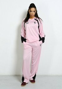 Młoda kobieta stoi z rękami w kieszeniach, ubrana w różowo-czarny sportowy strój i bordowe buty, na tle białej ściany.