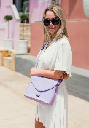 Levandulová syntetická crossbody taška s trojúhelníkovým klopem, zipovým uzávěrem a logem. Nositelka má dlouhé vlasy a je oblečená v lehkých bílých šatech.