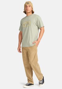 T-shirt de algodón verde claro con un parche de logo rectangular en amarillo y marrón, combinado con pantalones de pana beige y zapatillas negras.
