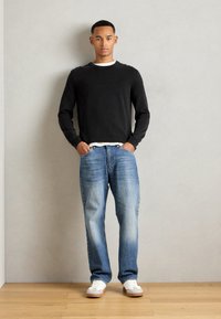 Junger Mann, der an einer hellen Wand steht, trägt ein schwarzes Langarmshirt, blaue Jeans, weiße Sneakers, die Hände in den Taschen, mit neutralem Ausdruck.