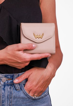 Beige textured wallet with a flap closure and a gold wavy logo. The wallet is being held by a hand against a black top and blue jeans.