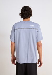 Šviesiai mėlynas trumpomis rankovėmis T-shirt, pagamintas iš medvilnės, su baltu logotipu ant viršutinės nugaros ir juoda raštuota juosta per petį.