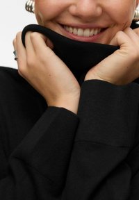 Czarny sweter dzianinowy z wysokim kołnierzem, prezentowany z dłońmi trzymającymi materiał blisko twarzy, podkreślający prążkowaną teksturę i rękawy.