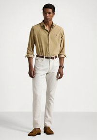 Koszula na guziki w kolorze beżowym z podwiniętymi rękawami, kremowe spodnie oraz brązowe skórzane buty. Na koszuli znajduje się małe logo, a do tego brązowy pasek.