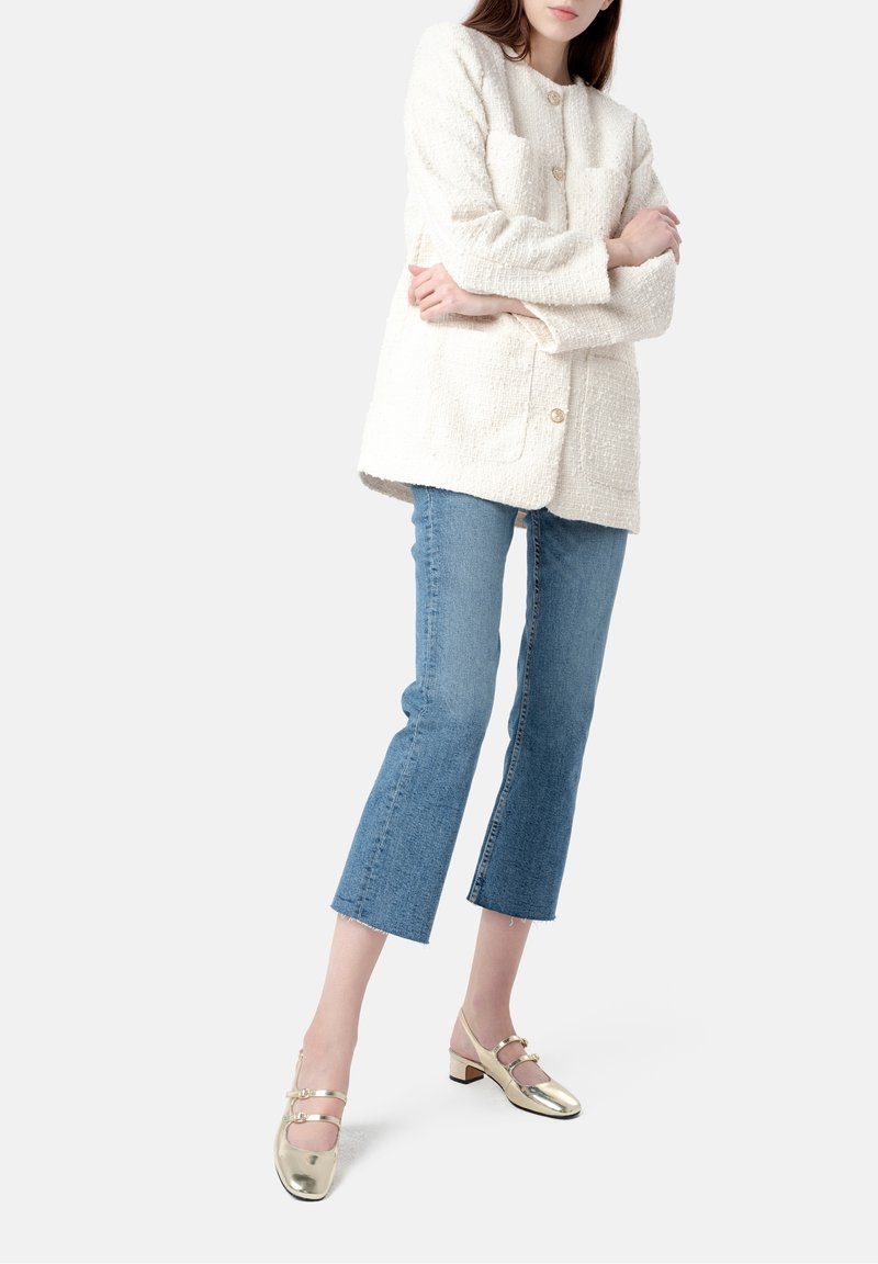 Femme portant un manteau texturé blanc, un jean bleu court et des chaussures Mary Jane dorées métalliques à talon bas, debout les bras croisés.