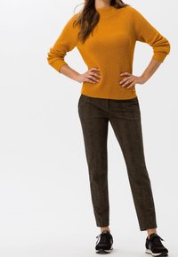 Femme portant un pull côtelé jaune moutarde, un pantalon slim brun foncé et des baskets noires, debout les mains sur les hanches devant un fond blanc.