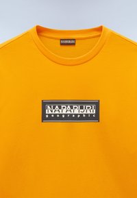 Keltainen puuvillapaita pyöreällä kaula-aukolla, jossa on mustavalkoinen suorakaiteen muotoinen logopainatus, jossa lukee "NAPAPIJRI geographic."
