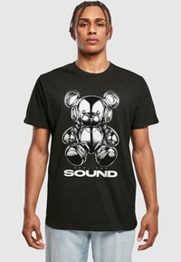 Svart t-skjorte med et trykk av en metallisk kosedyr med hodetelefoner, ledsaget av teksten "FEEL THE SOUND" i hvitt.