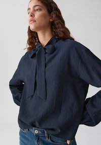 Blusa plissettata blu navy con cravatta, caratterizzata da maniche lunghe e una vestibilità comoda. Il tessuto ha una lieve texture e una finitura morbida.