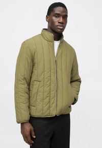 Casaco acolchoado verde-oliva com gola alta, fecho frontal com fecho éclair e dois bolsos laterais. Apresenta costura vertical e punhos elásticos.