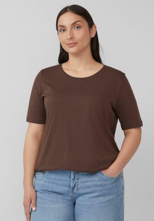 Bruine korte mouwen t-shirt met een ronde halslijn, gemaakt van een soepele stof. Gecombineerd met lichtblauwe jeans, handen in de zakken.