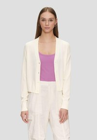 Cremfarbener Cardigan mit geripptem Kragen und fünf Knöpfen, über einem rosa gerippten Tanktop getragen. Das Model trägt hellfarbene, locker sitzende Hose.