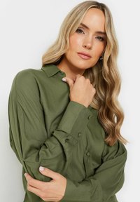 Chemise boutonnée vert foncé avec col, manches longues et texture lisse. Comprend des boutons sur les poignets et une coupe décontractée.