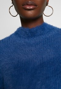 Pull bleu duveteux à col montant. Présente une finition douce et texturée. De grandes boucles d'oreilles dorées complètent la tenue.