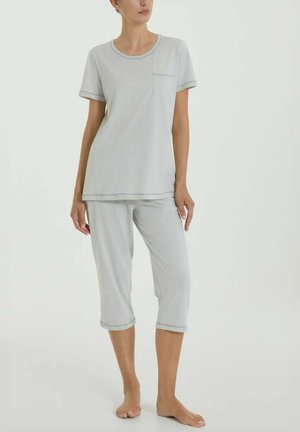 Donna che indossa una maglia a maniche corte color grigio chiaro con tasca e pantaloni corti coordinati, in piedi scalza davanti a uno sfondo semplice.