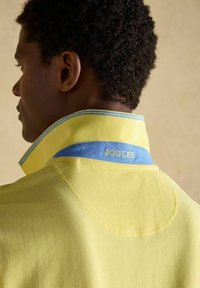 Homme aux cheveux courts et bouclés portant une chemise jaune à col avec un col intérieur bleu brodé de "JOULES" dans le dos.