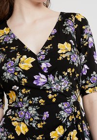 Schwarzes Kleid mit lebendigen gelben, lila und grünen Blumenmustern. V-Ausschnitt-Design mit kurzen Ärmeln und Wickeldetails an der Taille.