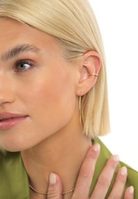 Auksinės spalvos kūgio formos auskarai su elegantišku, pailgu dizainu ir blizgiu paviršiumi. Modelis taip pat dėvi subtilią auksinę grandinėlės vėrinį.