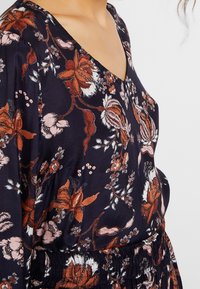 Dunkelblaues Kleid mit roten, rosa und weißen Blumenmustern, das über einen V-Ausschnitt und eine geraffte Taillendetails verfügt.