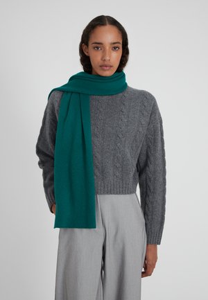 Pull en tricot gris associé à une écharpe teal, avec une texture douce. L'écharpe tombe élégamment sur les épaules. Pantalon gris clair.