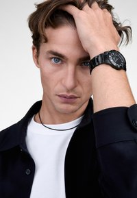 Sort ur med en rund urskive og linkarmbånd, båret på et håndled. Modellen har uordentligt hår og bærer en sort skjorte over en hvid t-shirt.