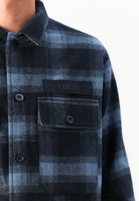 Uomo che indossa una camicia a quadrati blu scuro e nera con taschino e etichetta "REPLAY" sopra il taschino.
