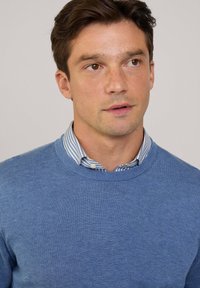 Blauwe trui met een ronde hals, gemaakt van een gestructureerde brei. Gedragen over een witte blouse met kraag met blauwe strepen.