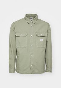Green cotton shirt with a collared neck, two chest pockets, and silver snap buttons. Features a small label on the left pocket.