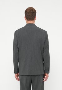 Bărbat văzut din spate, îmbrăcat într-o jachetă de costum gri cu dungi subțiri și pantaloni asortați, pe un fundal simplu de culoare deschisă.