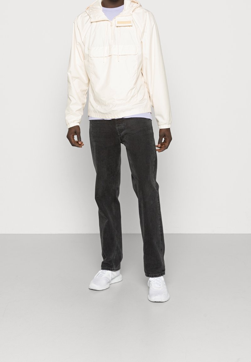Personne portant une veste à capuche beige clair, un jean noir et des baskets blanches, se tenant devant un fond uni de couleur claire.