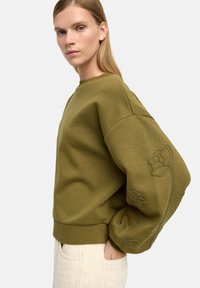 Oliwkowy sweatshirt z oversize'owymi rękawami, ozdobiony haftowanymi kwiatami na lewym rękawie oraz ściągaczem przy dekolcie i u dołu.