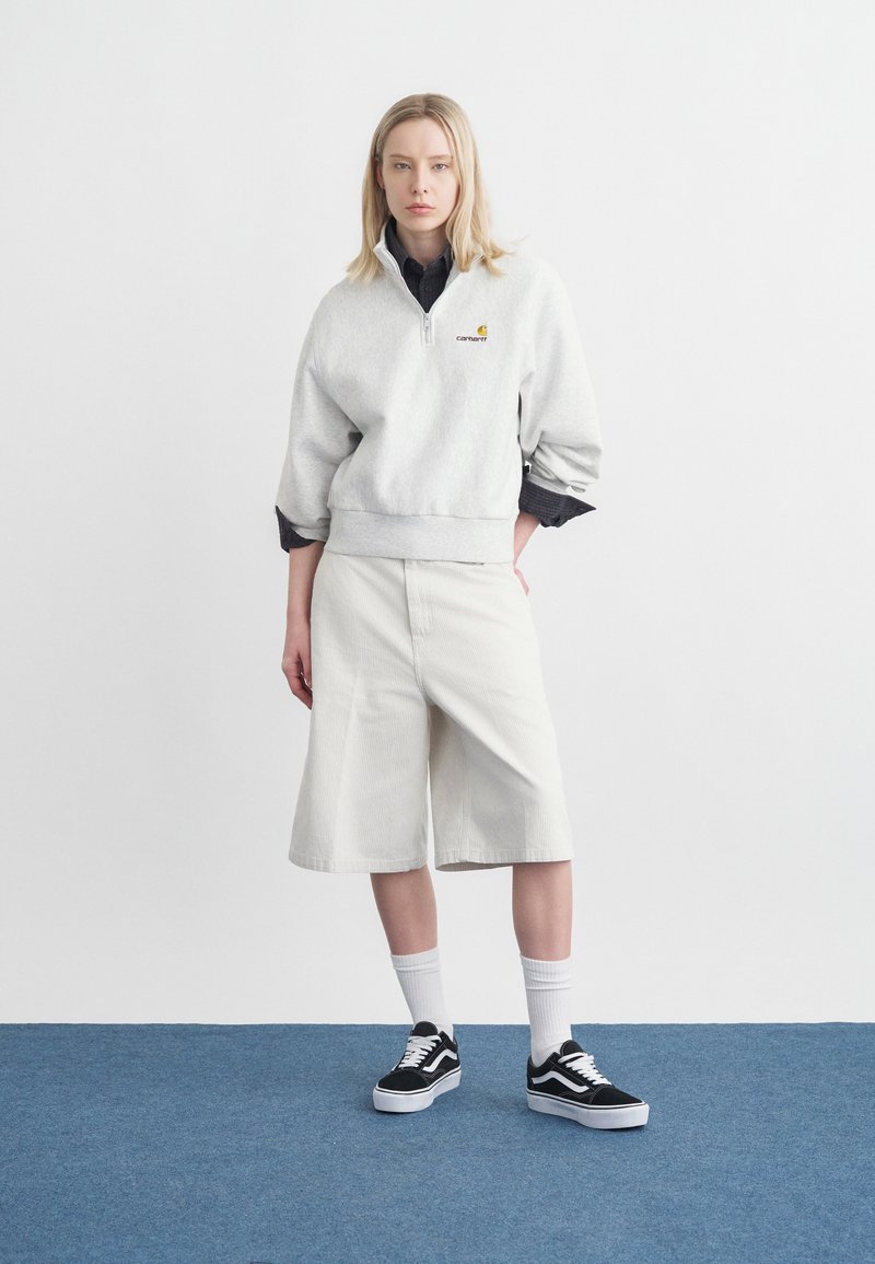 Donna bionda in piedi su un tappeto blu che indossa una felpa bianca con zip al quarto, pantaloncini bianchi fino al ginocchio, maglietta nera, calzini bianchi e scarpe da ginnastica nere.