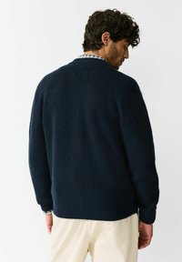 Pulover tricotat bleumarin cu textură nervurată, guler rotund și umeri căzuți, peste o cămașă în carouri, asortat cu pantaloni de culoare crem.