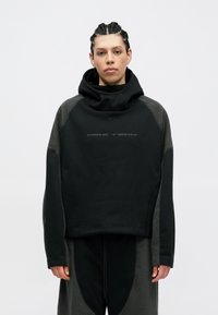 Fekete Nike Tech kapucnis pulóver magas gallérral, szürke bolyhos részletekkel a vállakon és az oldalsó panelekben. Texturált anyag és laza szabás.