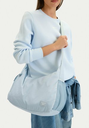 Person, die ein hellblaues Langarmshirt trägt, eine hellblaue Reebok-Schultertasche mit sich führt und eine Jeansjacke hält.