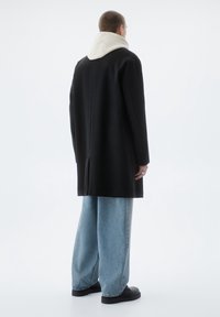 Cappotto in lana nero con colletto in felpa bianca, vestibilità ampia e jeans a taglio dritto. Presenta un design minimalista e una texture sottile.