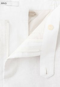Tissu blanc en gros plan montrant une ceinture avec un bouton et une boutonnière, une doublure intérieure et une petite étiquette en tissu avec le texte "MNG".