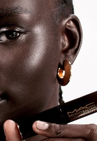 Svart tub med MAC Studio Radiance foundation som hålls i en hand, nära ett ansikte med slät, mörk hy och ett stort guldörhänge.