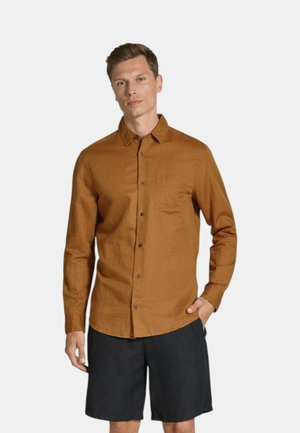Camicia marrone button-up in un tessuto leggero, con taschino sul petto e orlo arrotondato, abbinata a pantaloni corti scuri.
