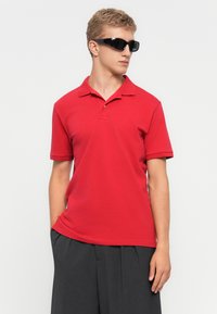 Junger Mann mit schwarzer Sonnenbrille, rotem Poloshirt und dunkelgrauen Hosen, der mit einer Hand in der Tasche vor einem weißen Hintergrund steht.