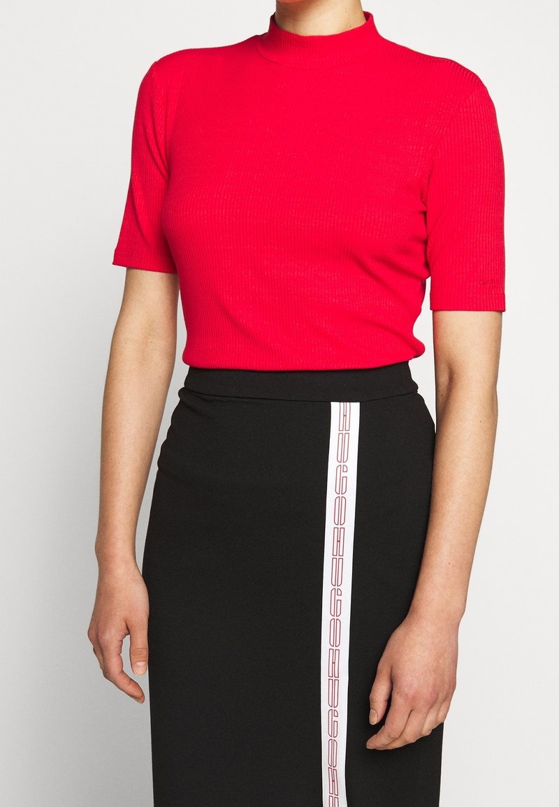 Femme portant un haut rouge vif à manches courtes et une jupe noire taille haute avec une bande blanche verticale sur le devant.