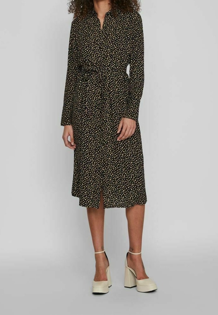 Femme portant une robe midi noire à pois blancs, manches longues et chaussures à talons hauts compensés crème sur un fond uni.