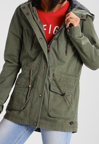 Person som bär en olivgrön jacka med flera fickor och dragsko, delvis öppen för att visa en röd tröja under.