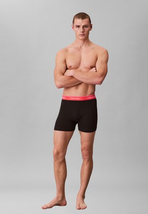 Spierbundige jonge man staat blootsvoets met gekruiste armen, draagt zwarte boxer briefs met een rode Calvin Klein tailleband.