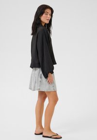Top corto nero e texturizzato con maniche larghe, abbinato a una gonna di jeans grigio chiaro. Modella che indossa infradito nere, in piedi di lato.