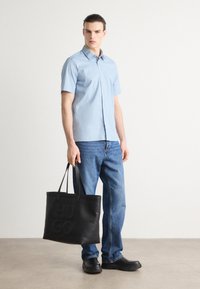 Világoskék, rövid ujjú ing, sötétkék farmernadrág, fekete táska dombornyomott "HUGO" felirattal, és fekete slip-on cipők egy semleges háttér előtt.