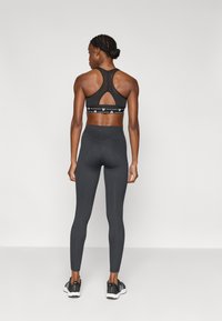 Μαύρο αθλητικό σουτιέν με πίσω άνοιγμα και διακοσμητική ζώνη κάτω, σε συνδυασμό με σκουρόχρωμο γκρι ριμπ κολάν. Το μοντέλο φοράει μαύρα αθλητικά παπούτσια.